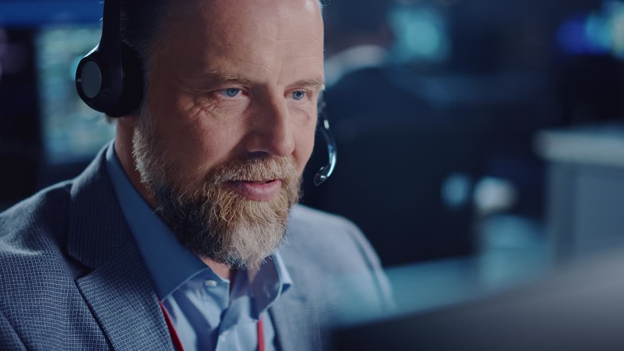 Close Up Portrait of a Joyful Bearded Technical Customer Support Specialist Talking on a Headset while Working on a Computer in a Call Center Room Filled with Computer Display Screens and Data Servers