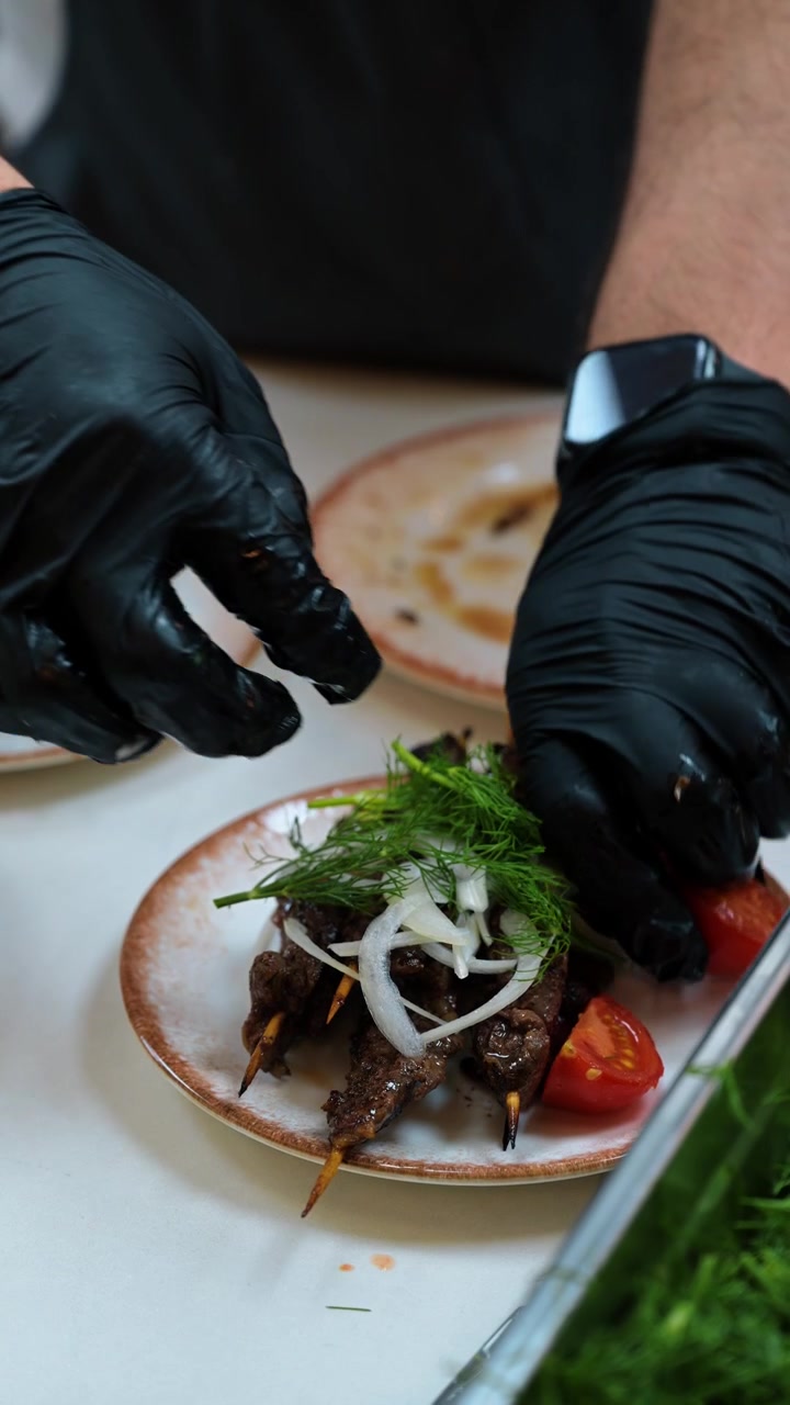 Gloved hand garnishing meat skewers. Dill and sliced onion top browned pieces on wooden skewers in a close kitchen view.