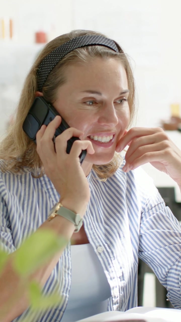 Vídeo vertical: Mulher de camisa listrada atendendo uma chamada, segurando um telefone preto e verificando um caderno em casa.