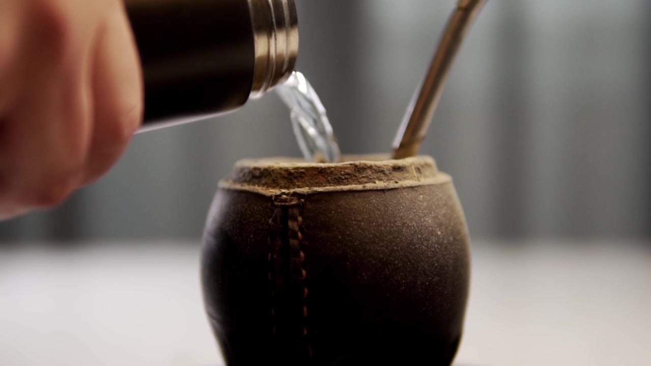 Close view of water pouring into traditional herbal drink vessel with metal straw and dried herbs inside. Rustic ceramic container sits against soft background