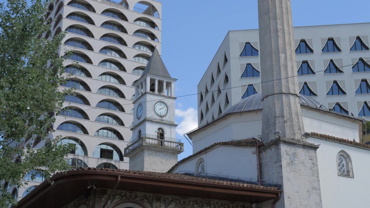 Minarete e torre do relógio da Mesquita Et'hem Bej erguem-se em frente a edifícios de hotel modernos e padronizados no centro de Tirana. Plano inclinado para baixo.