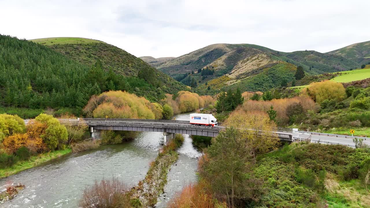 A vehicle crosses a bridge surrounded by lush greenery and hills under soft daylight at Lake Tekapo