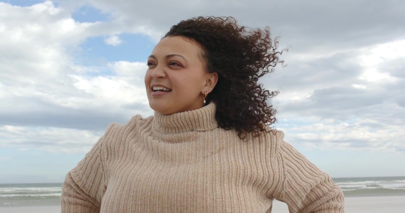 Donna afroamericana in piedi sulla spiaggia che indossa un maglione, con l'oceano sullo sfondo, che reagisce al vento che si alza, ridendo.