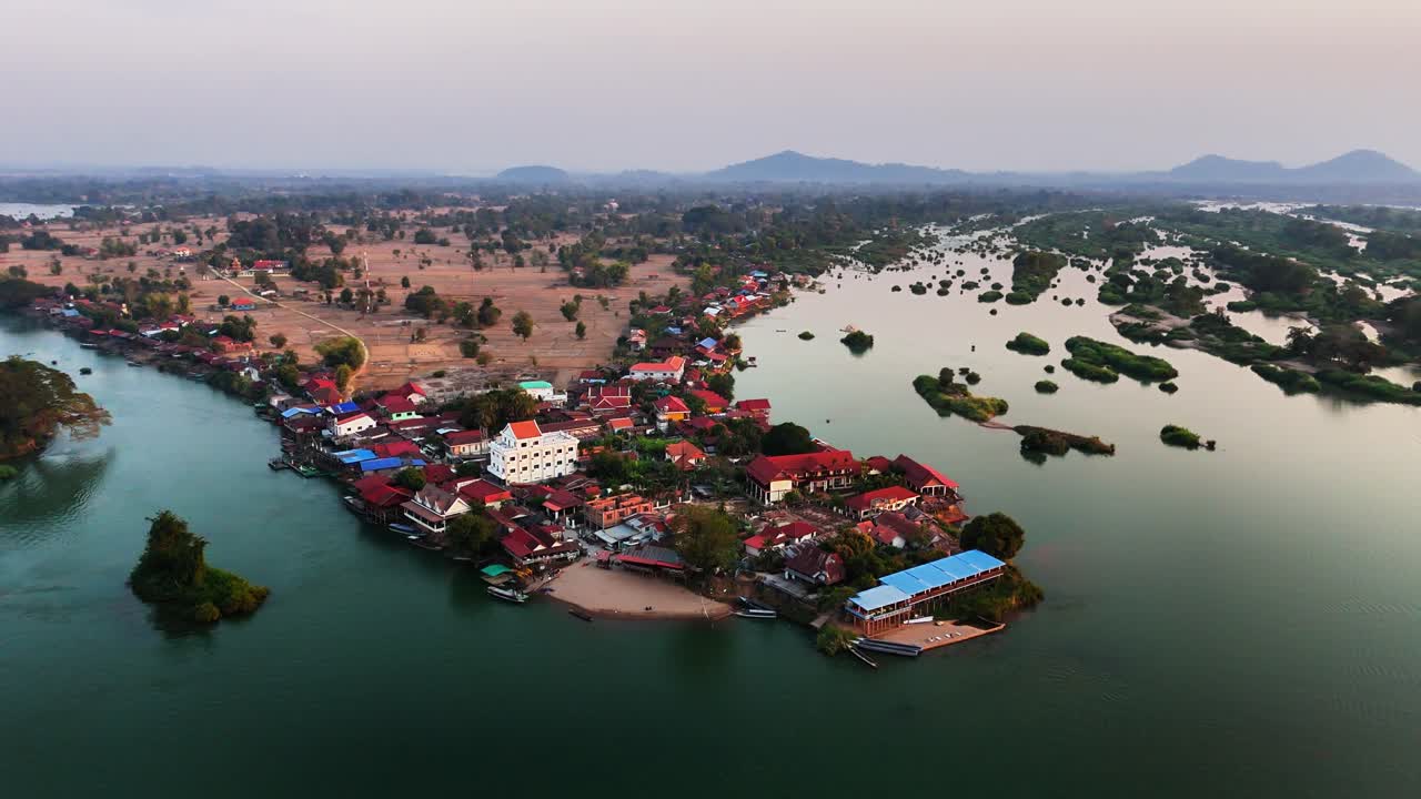 Aerial drone orbit reveal core of Don Det village in Laos, riverside stilted bungalows, historic wooden bridge linking to Don Khon, and the small Buddhist temple along the Mekong shoreline at sunset