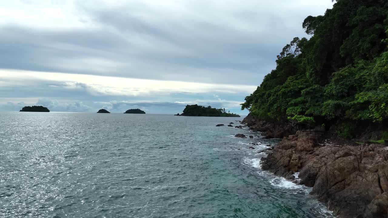 Low-angle drone flight gliding slowly along the rocky shoreline of Lonely Beach on Koh Chang Island, Trat, Thailand, capturing turquoise Gulf of Thailand waters and lush tropical greenery