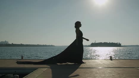beautiful woman in long black dress walks across wooden deck near the sea - static, wide shot