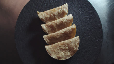 hermosa toma en cámara lenta de un grupo de 4 tacos o tortillas mexicanas mientras la cámara se desplaza y hace zoom alrededor del plato negro y las tortillas iluminadas con una suave luz blanca