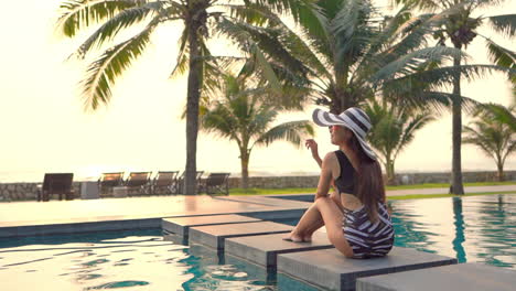 woman with swimsuit and large hat sitting on pool edge and looking at panorama