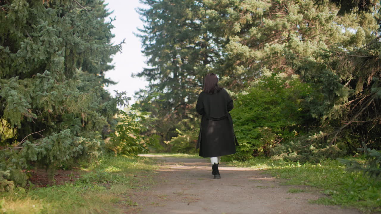 Вид со спины на молодую девушку в повседневной одежде и длинном зимнем пальто, прогуливающуюся по дендрарию с телефоном в руке и снимающую природу под соснами во время осенней прогулки.