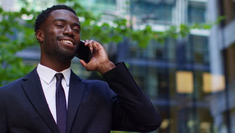 Smiling-Young-Businessman-Wearing-Suit-Talking-On-Mobile-Phone-Standing-Outside-Offices-In-The-Financial-District-Of-The-City-Of-London-UK