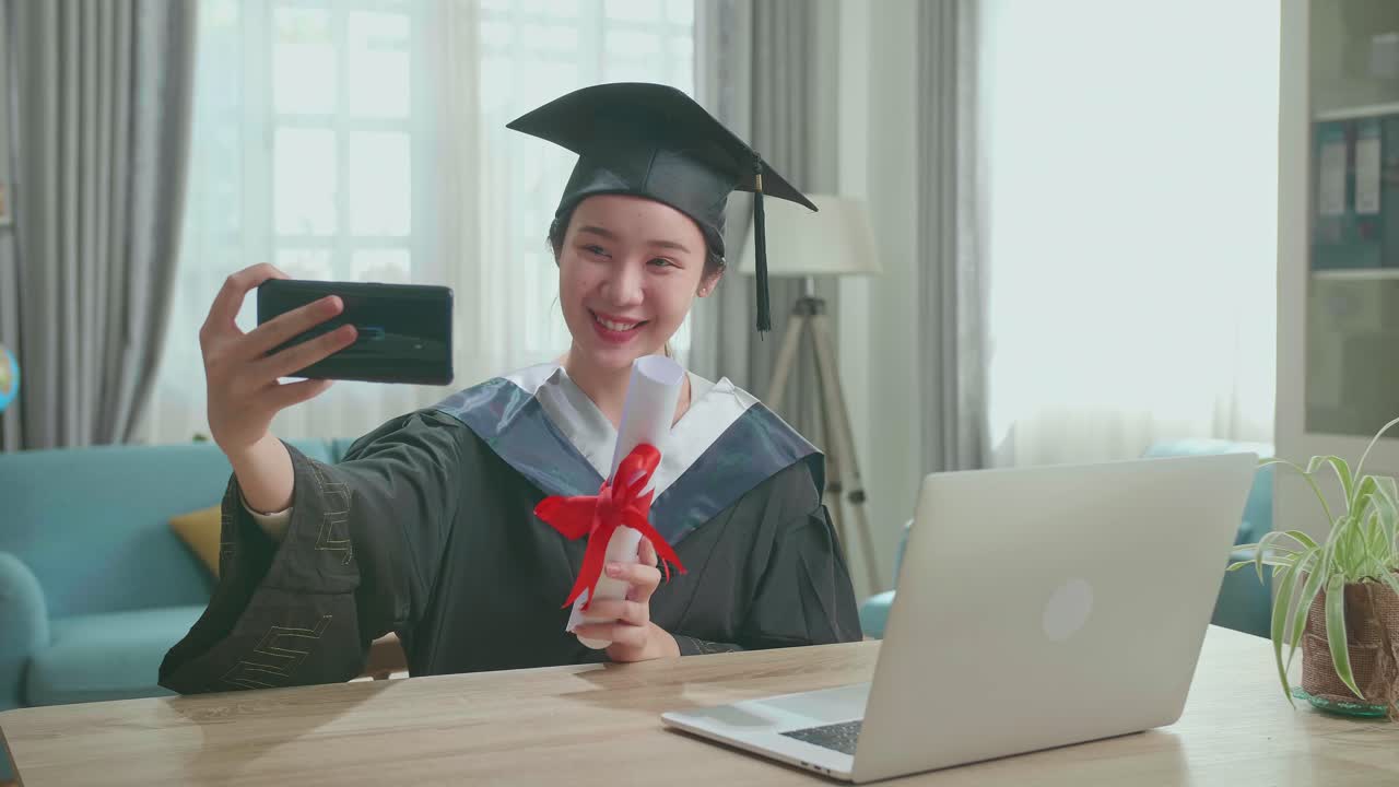 Young Asian Woman Taking A Selfie While Wearing A Graduation Gown And Cap, Hand Holding University Certificate In Living Room