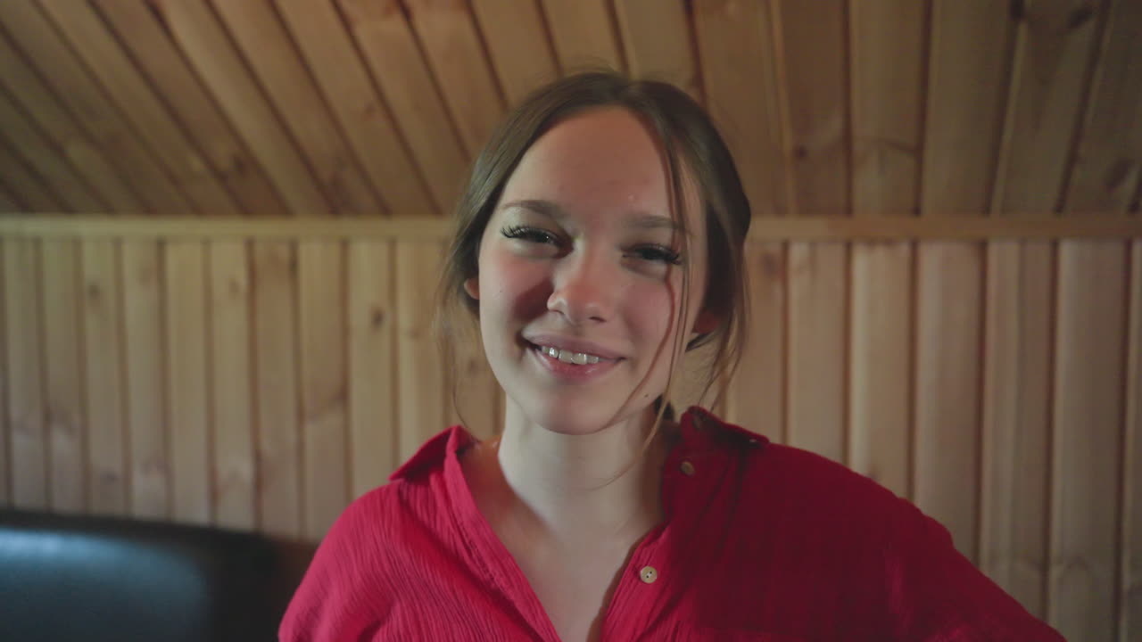 beautiful young woman standing indoors smiling softly toward camera, dressed in red shirt with natural expression, wooden wall background