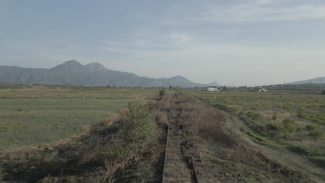 Drone-shot-that-follows-old-and-unused-train-track-in-the-valley-between-the-mountains-in-Albania-on-a-cloudy-day-LOG