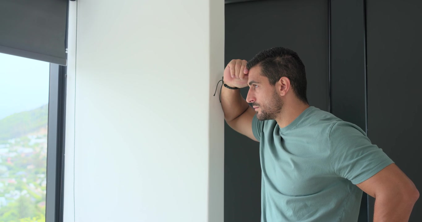 Uomo adulto appoggiato a una parete bianca alla finestra di casa, che guarda fuori e poi si strofina la fronte, con spazio per il testo.