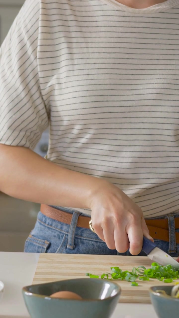 Video verticale: Una donna in maglietta a righe affetta cipollotti su un tagliere in cucina per la preparazione dei pasti.