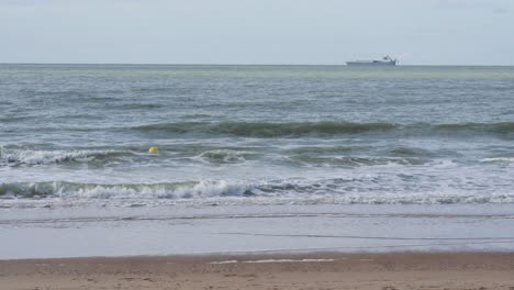 A-single-yellow-buoy-bobs-in-gentle-waves-as-an-ocean-going-ship-slowly-passes,-North-Sea,-Belgian-coast
