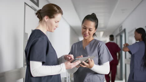 Diverse-female-doctors-discussing-work,-using-tablet-in-corridor-at-hospital,-slow-motion