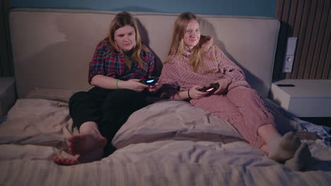 teenage girls playing video games in pajamas on a bed at night