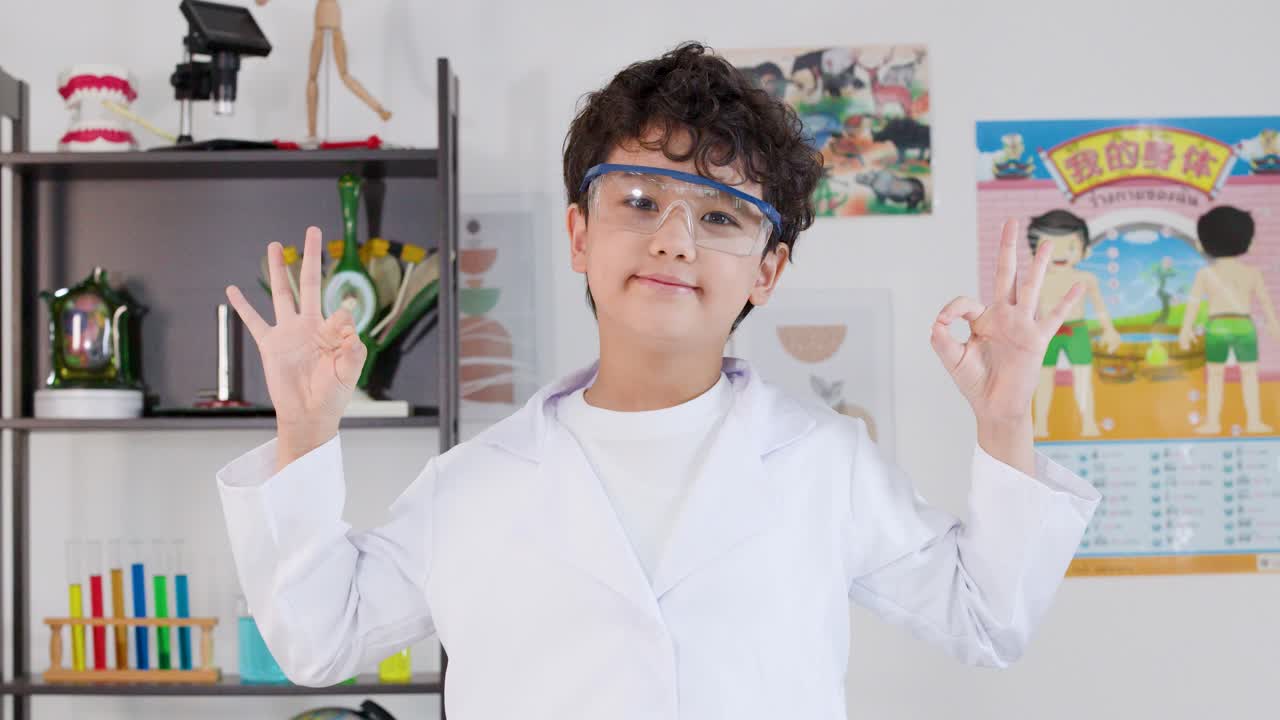 白い白衣と安全ゴーグルを着用したアジア系の学生が、屋内で手のジェスチャーをしている。
