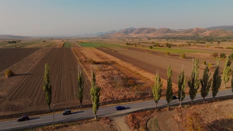 Drohnenverfolgung,-Weitwinkelaufnahme,-Blaues-Auto,-Das-An-Einem-Sonnigen-Tag-Auf-Einer-Sonnenbeschienenen,-Gepflasterten-Straße-In-Richtung-Berge-Fährt,-Umgeben-Von-Grünen-Bäumen