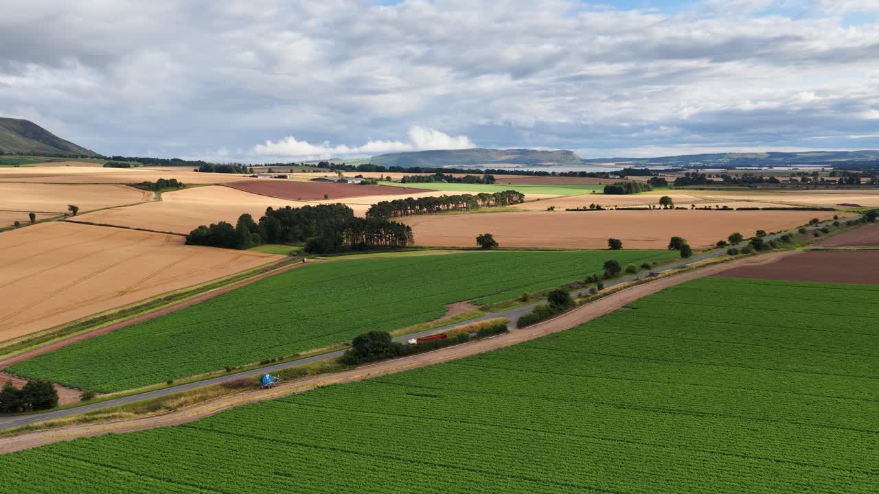 Aerial camera smoothly pans across patchwork fields, green crops, and golden harvest under cloudy daylight, revealing expansive rural landscape near Kinross, Scotland