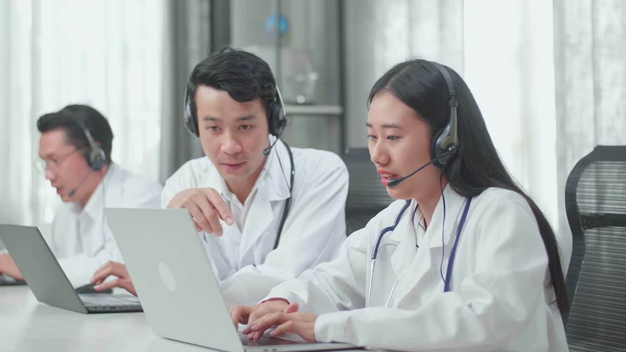 A Man And A Woman Of Three Asian Doctors With Stethoscopes In Headsets Working As Call Center Agent Are Discussing Work While Their Colleague Is Speaking With Customer On The Call At The Office