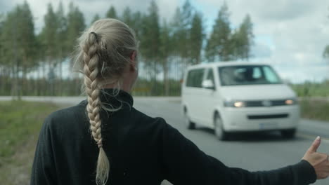 Hermosa-Chica-Rubia-Haciendo-Autostop-En-Un-Cruce-De-Caminos,-Campo-De-Finlandia,-Parada-Automática-Femenina-Con-Mochila-Pesada-En-Escandinavia,-Los-Autos-No-Se-Detienen,-Primer-Plano-Desde-Atrás