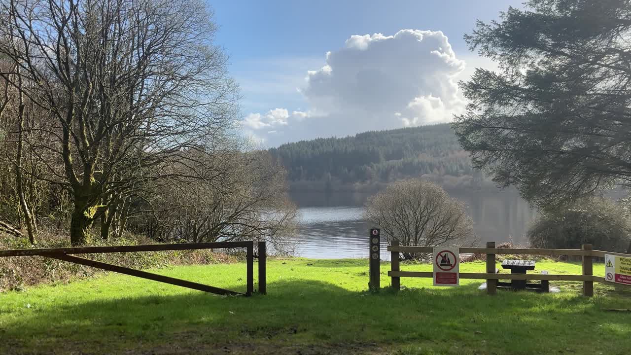 Breedbeeldopname van een vredig veld aan het meer op het platteland van Wales, omlijst door bomen en houten hekken, met kalm water en beboste heuvels onder zacht middaglicht en drijvende wolken
