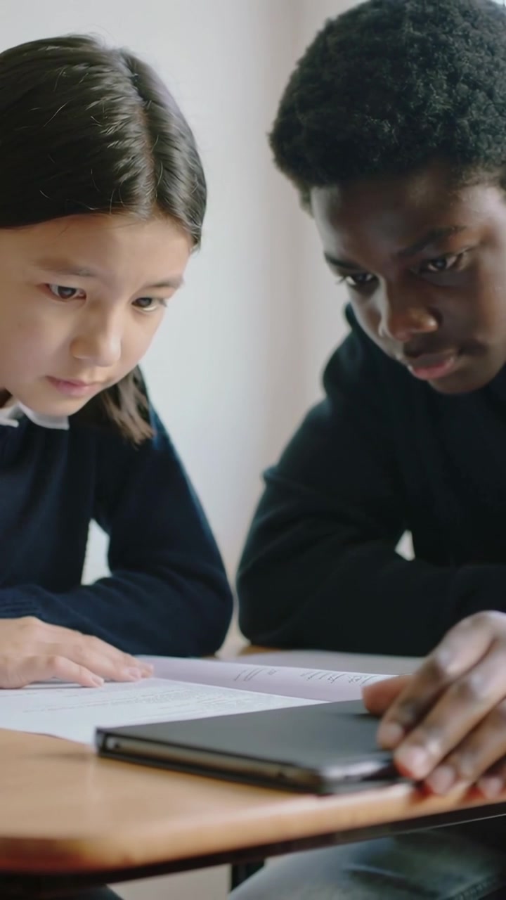 Video verticale: Studenti chinati con maglioni scuri che leggono un quaderno di esercizi al banco di scuola, facendo scorrere un tablet.
