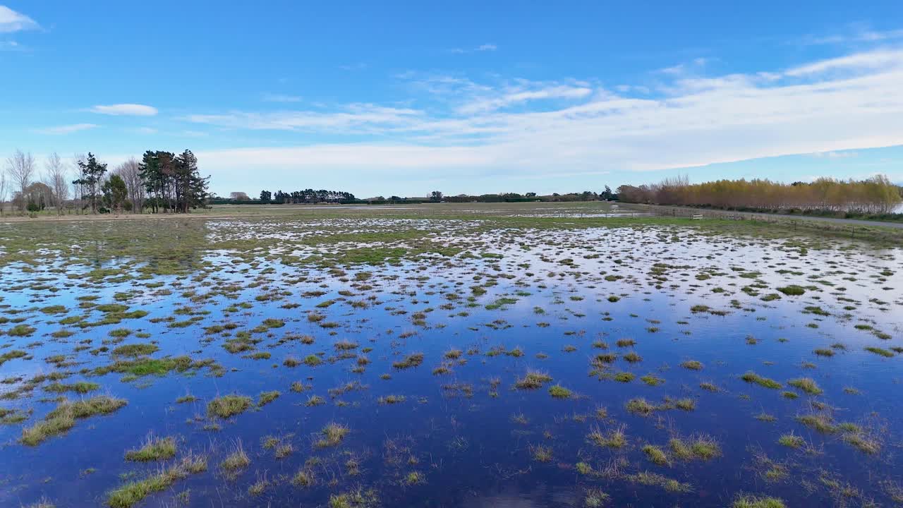Aerial view of a flooded field under a clear blue sky, showcasing water reflections and sparse vegetation