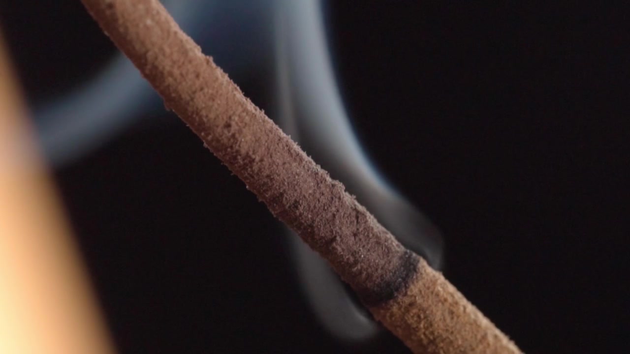 Macro ravvicinata di un bastoncino d'incenso che brucia con fumo che sale su sfondo nero. Vista dettagliata dell'incenso incandescente che rilascia fumo bianco sottile con superficie testurizzata.