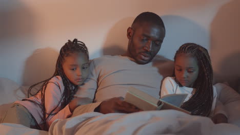 father reading book to daughters in bed
