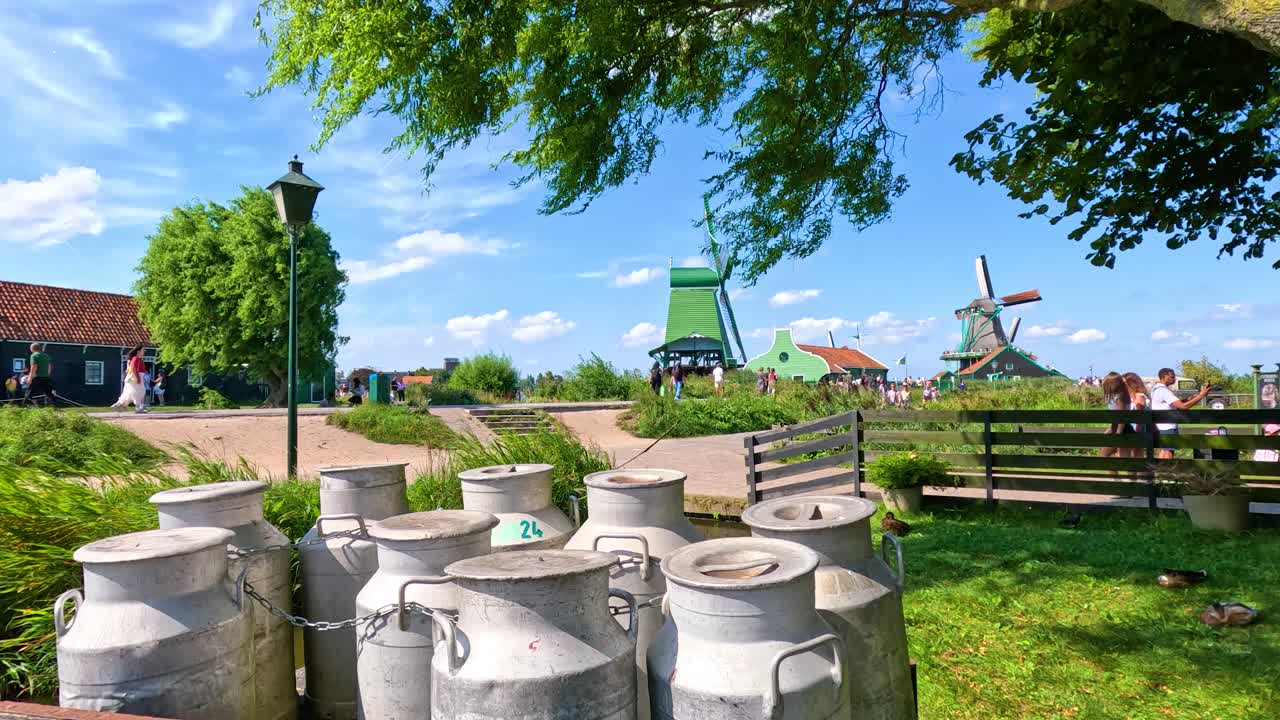 Camera pans across windmills, milk cans, and tourists in a sunny, vibrant Dutch countryside setting