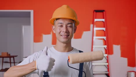 primer plano de un pintor asiático que usa un casco de seguridad sonriendo y mostrando los pulgares hacia arriba mientras está de pie con la pared de pintura en la habitación