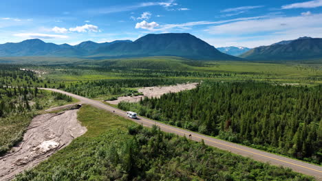 white lonely campervan moving by the highway in the wilderness of alaska. aerial perspective on the vast valley with mountain range at backdrop