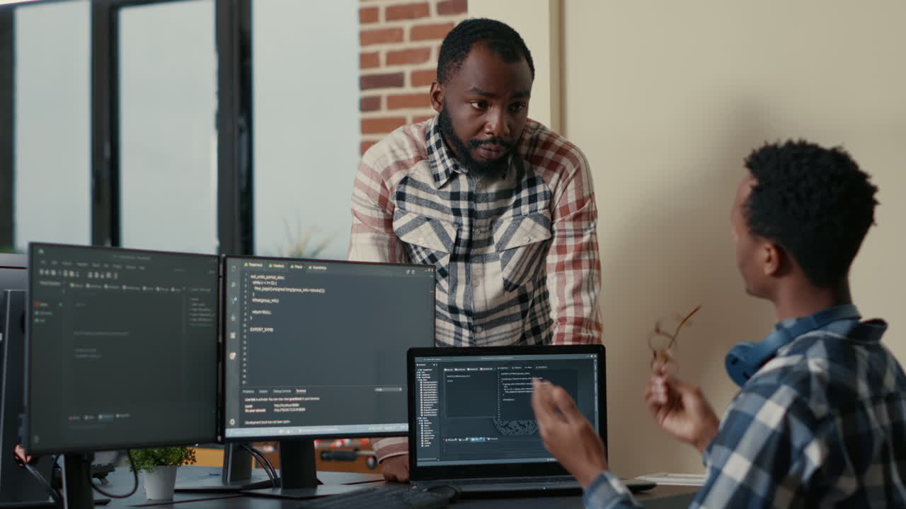 Software developer sitting at desk with multiple screens and laptop running code talking with colleague