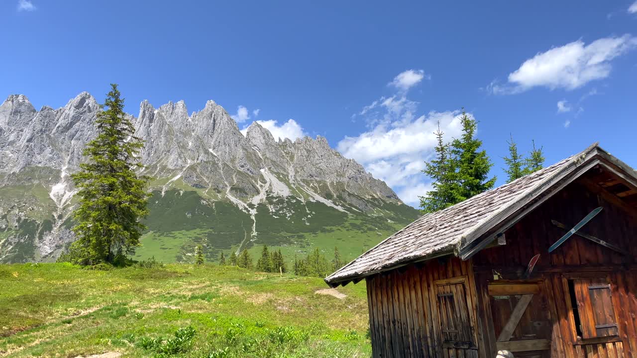 Ujęcie panoramiczne starego drewnianego domku turystycznego w zielonym krajobrazie ze skalistymi górami w tle.