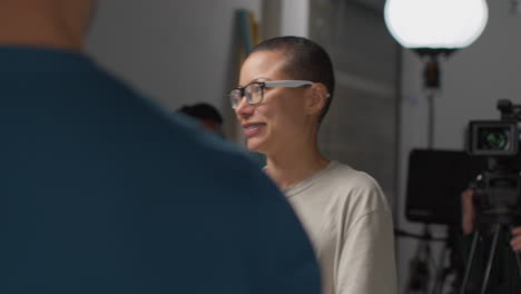 a woman on a film set, holding a clipboard, smiling and talking to someone off camera