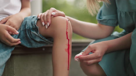 a close-up of a woman carefully cleaning a child's injured knee with tissue paper, removing blood after a fall