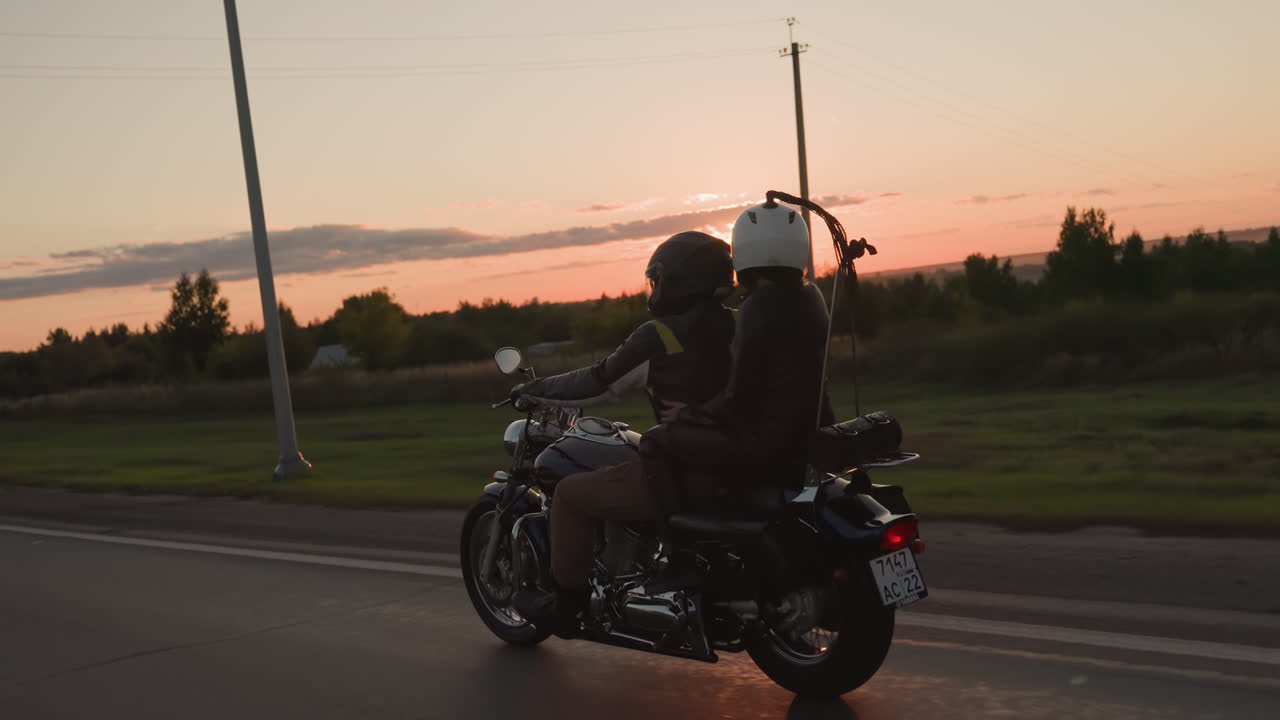 Couple wearing helmets rides motorcycle along highway during sunset, passing green fields and streetlights while traveling on road trip, symbolizing freedom, and adventure lifestyle on open road