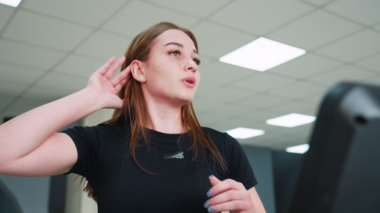 exhausted woman in black workout outfit wipes sweat from face during fitness session inside bright spacious gym with white ceiling panels and overhead lights in background