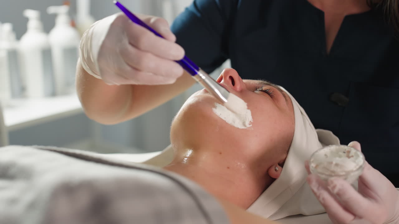 Skin Expert Applies Thick White Face Mask With Silicone Brush From Small Transparent Bowl Onto Relaxed Female Client Wrapped In White Towel During Professional Spa Facial Treatment Under Lighting