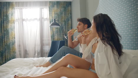Cute-couple-toasting-glasses-in-apartment.-Charming-couple-sitting-on-hotel-bed.