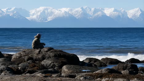a person sits on rocks by the ocean with majestic snow-capped mountains in the distance