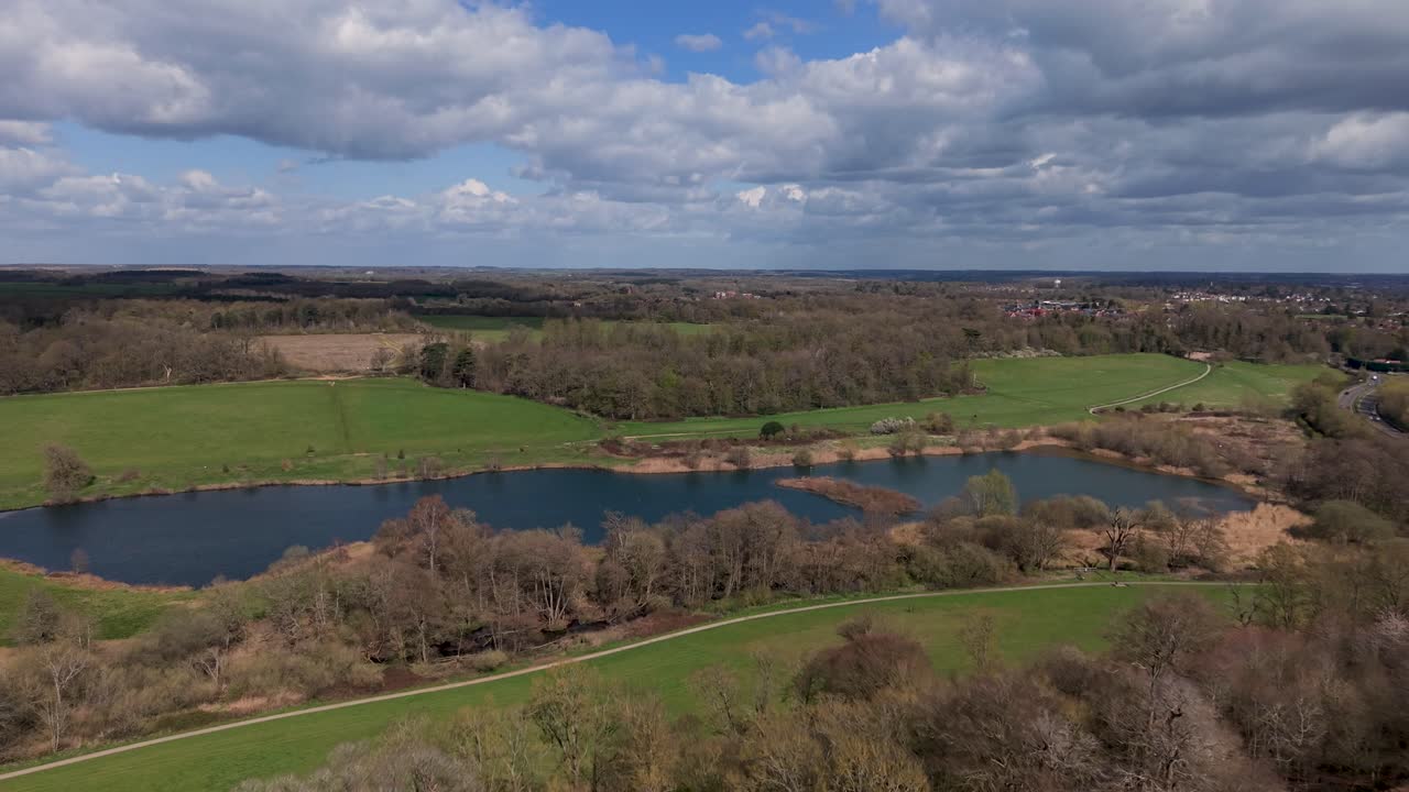 Volo di drone sopra il lago Osprey, Panshanger Park nell'Hertfordshire, Inghilterra. Planata aerea in avanti sopra acqua calma, boschi e campi verdi sotto luminose nuvole primaverili velate.