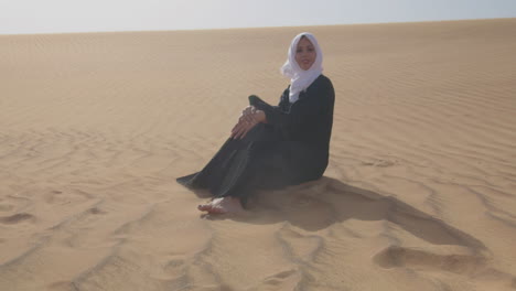 Beautiful-Muslim-Woman-In-White-Hijab-And-Traditional-Black-Dress-Sitting-On-Sand-In-A-Windy-Desert