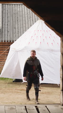 armed man in cloak walks near wooden gate and stops to watch territory carefully. serious warrior stands near white tent in courtyard of medieval castle