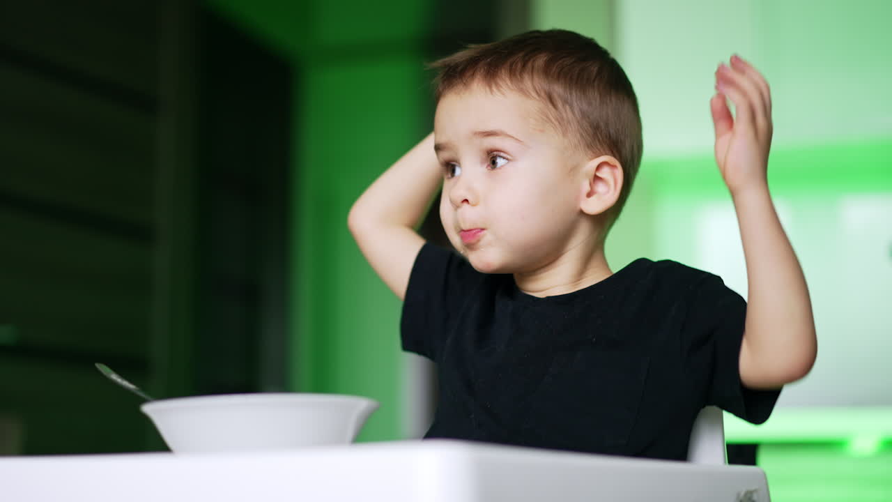 Adorabile bambino seduto davanti al piatto. Il bambino agita le mani allegramente durante i pasti. Vista dal basso. Sfondo sfocato.