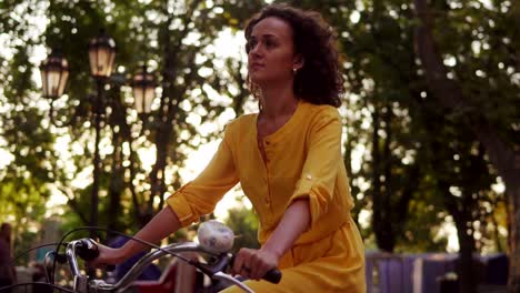 lens flare: young woman in long yellow dress is riding a city bicycle with a basket and flowers in the park with green trees