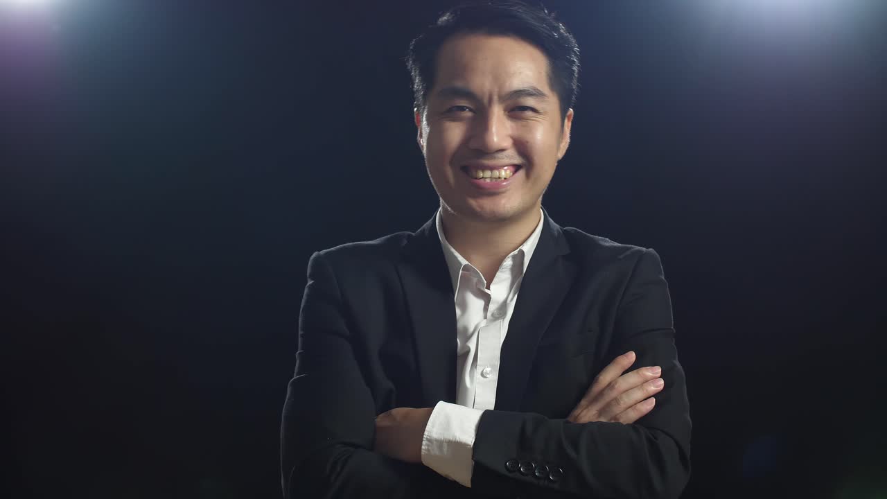 ブラックスクリーンスタジオに立っている間に腕を交差するビジネススーツを着た笑顔のアジア人スピーカーの男性